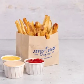 A brown paper bag of French fries labeled "Jeff's Beach Burgers" sits on a gray surface next to three dipping sauces in small white cups: ketchup, cheese, and a creamy sauce.