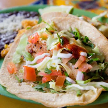 Two fish tacos topped with lettuce, tomato, and sauce are served on a green plate with a side of black beans and rice.