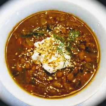 A bowl of thick vegetable stew topped with a dollop of cream, sprinkled nuts, and a sprig of fresh dill.