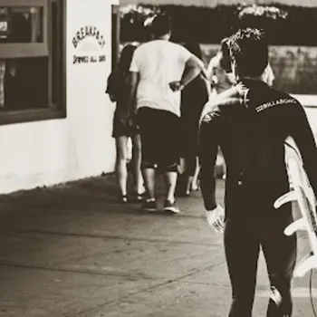 A person in a wetsuit carries a surfboard while walking on a sidewalk toward a group of people near a building.