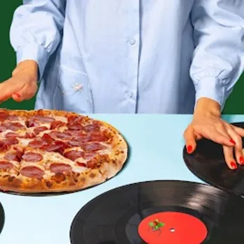 A person in a blue shirt stands behind a table, touching a pepperoni pizza and two vinyl records arranged side by side.