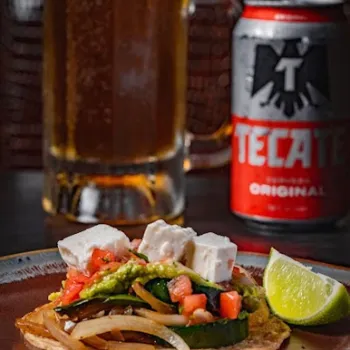 A plate with a tostada topped with vegetables and cheese, a lime wedge, a glass of beer, and a can of Tecate beer in the background.