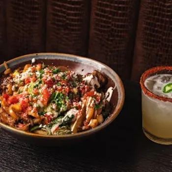 A bowl of loaded fries topped with tomatoes, cheese, and sauce sits next to a cocktail garnished with lime, jalapeño, and a spiced rim on a dark table.