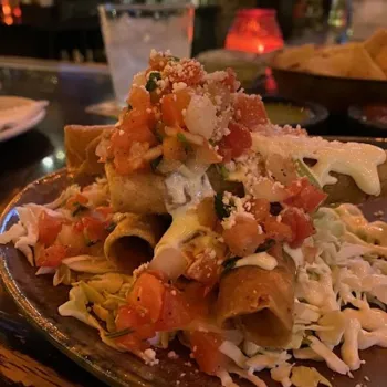 A plate of taquitos topped with diced tomatoes, onions, cilantro, crema, and cheese, served on a bed of shredded cabbage in a dimly lit restaurant.