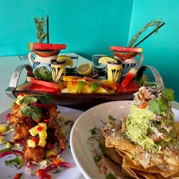 Two cocktails with fruit garnish sit behind plates of fried chicken with mango salsa and stacked nachos topped with guacamole, against a turquoise wall.