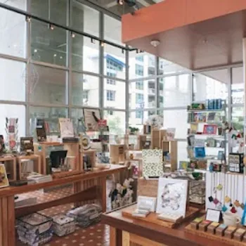 A well-lit gift shop with wooden tables and shelves displaying books, art prints, postcards, and small gift items, set against large windows with an urban view.