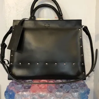 A black leather handbag with silver studs, a cross symbol, and a shoulder strap, displayed on a stand made of pink and blue bubble packaging.