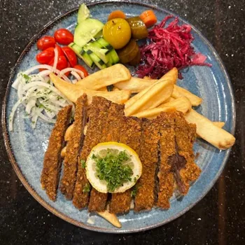 A plate with breaded meat cutlets, fries, a lemon slice, and sides of cucumber, cherry tomatoes, onions, olives, pickles, and shredded red cabbage.