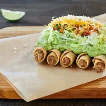 Five rolled taquitos topped with guacamole, shredded lettuce, diced tomatoes, and shredded cheese on parchment paper and a wooden board, with a lime wedge in the background.