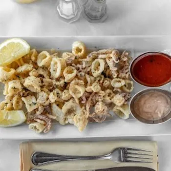 A rectangular white plate with fried calamari, two lemon wedges, cocktail sauce, and a creamy dip, set on a white tablecloth with a fork and knife on a napkin.