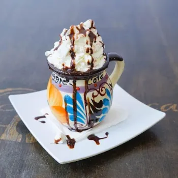 A ceramic mug with colorful patterns is filled with hot chocolate, topped with whipped cream and chocolate syrup, placed on a white square plate with some spillage.