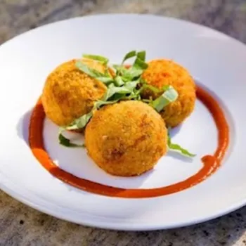 Three breaded and fried balls are served on a white plate with a ring of red sauce and garnished with green herbs.