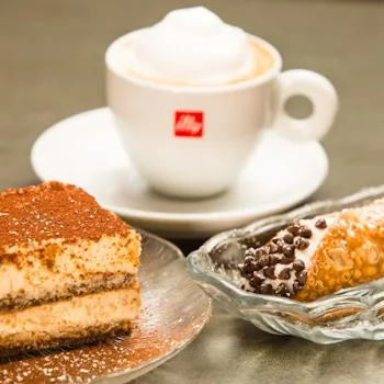 A cup of cappuccino on a saucer, with a slice of tiramisu on a glass plate and a chocolate chip cannoli on a glass dish.