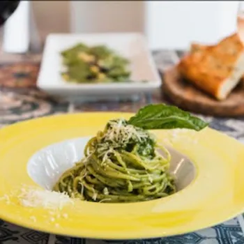 A plate of pesto spaghetti topped with basil and grated cheese, served with bread, salad, and glasses of red and rosé wine on a patterned table.