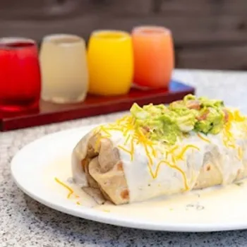 A burrito topped with white sauce, shredded cheese, and guacamole is served on a white plate, with four colorful drinks in small glasses in the background.