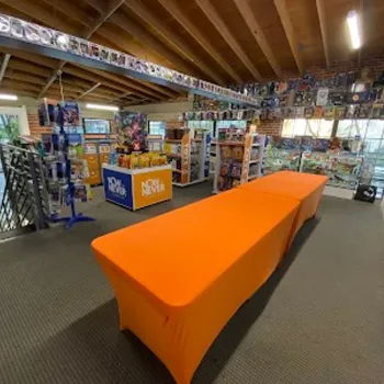 A game store interior with two orange-covered tables, display shelves filled with board games and toys, and large windows letting in natural light.