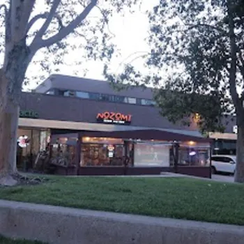 A small shopping plaza at dusk features Nozomi Japanese restaurant, a dental office, parked cars, trees, and a grassy area in the foreground.
