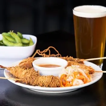 A plate with skewered fried noodles, dipping sauce, and pickled vegetables, with a bowl of edamame and a pint of beer on a dark table.