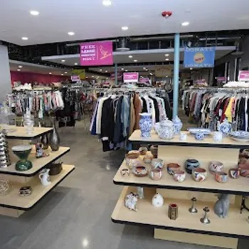 A thrift store interior with racks of clothing and shelves displaying various ceramic and glass items, all under bright lighting.