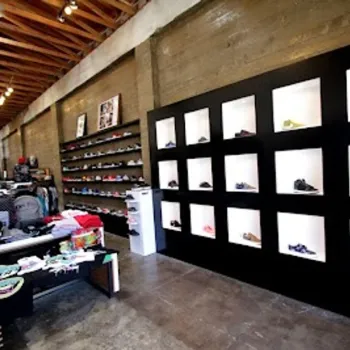 A modern sneaker store interior with shoes displayed in wall cubbies, shelves of footwear, and tables with accessories on a polished concrete floor.