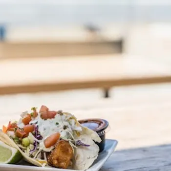 A plate with two fish tacos topped with slaw and diced tomatoes, garnished with a lime wedge, and accompanied by a small container of sauce on a wooden table outdoors.