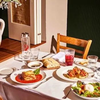 A table set for two with plates of Italian food, bread, salad, drinks, glasses, and utensils, next to a window and a large ceramic jug.