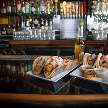 Two plates of tacos with dips, bottles of beer, and a bowl of nuts sit on a bar counter with liquor bottles and beer taps in the background.