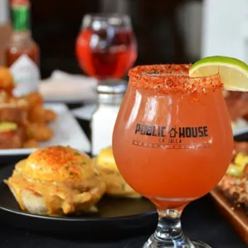 A glass of red-orange cocktail with a spiced rim and lime wedge, labeled "Public House La Jolla," sits on a table with food and other drinks in the background.