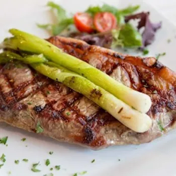 Grilled steak topped with two green onions, garnished with chopped herbs, served with a small salad of greens and cherry tomatoes on a white plate.