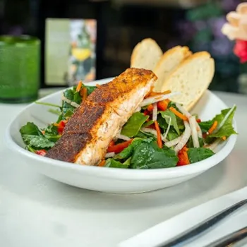A bowl of salad with leafy greens, sliced vegetables, a grilled salmon fillet, and two pieces of baguette on a white plate.