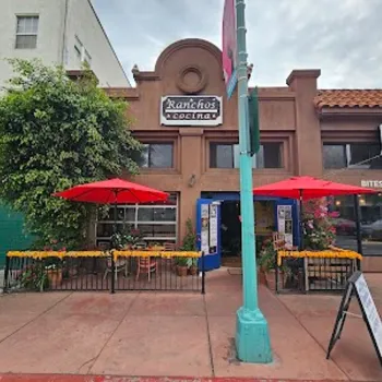 Street view of Rancho's Cocina restaurant with two red umbrellas over outdoor tables, marigold decorations on the patio railing, and a sign above the entrance.