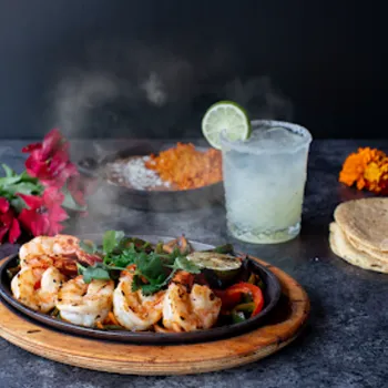 A sizzling plate of grilled shrimp and vegetables is served with tortillas, rice, a lime-garnished drink, and flowers on a dark surface. Steam rises from the food.