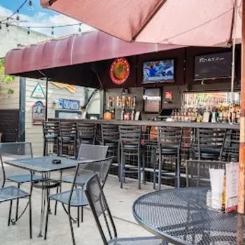 Outdoor bar patio with metal tables and chairs, overhead string lights, TVs above the bar, and a large umbrella providing shade.
