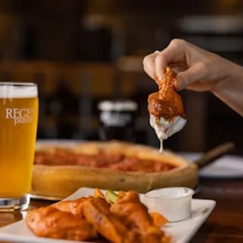 A hand dips a buffalo wing into ranch dressing, with a plate of wings, a glass of beer, and a pizza in the background on a wooden table.