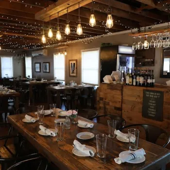 A rustic restaurant dining area with a wooden table set for eight, surrounded by chairs, hanging lights, and string lights overhead.