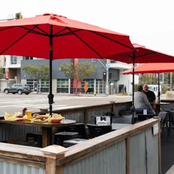 Outdoor restaurant seating area with red umbrellas, empty tables, and two people conversing at a table; food and drinks are on the tables, with a street and buildings in the background.