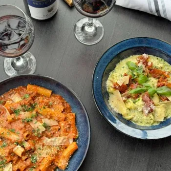Two plates of pasta—one with red sauce and rigatoni, the other with green sauce and shaved cheese—are served with two glasses of red wine on a dark table.