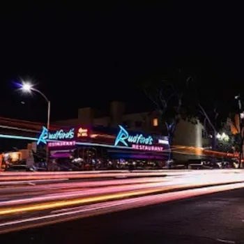 A neon-lit restaurant with "Rudford’s Restaurant" signage at night, with light trails from passing vehicles on the street in the foreground.