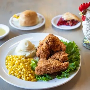 A plate with fried chicken, mashed potatoes, and corn. In the background, a dinner roll, a slice of pie, a small bowl of sauce, and a ceramic rooster are visible.