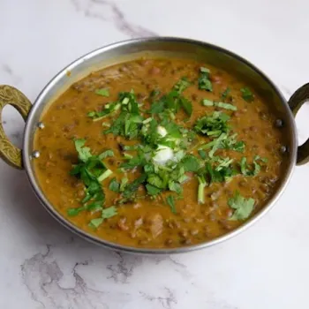 A bowl of dal makhani garnished with chopped cilantro and a dollop of cream, served in a metal dish on a marble surface.