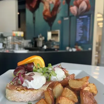 A plate of roasted potatoes and toast topped with poached eggs, microgreens, and an orange slice in a café with floral wall art in the background.
