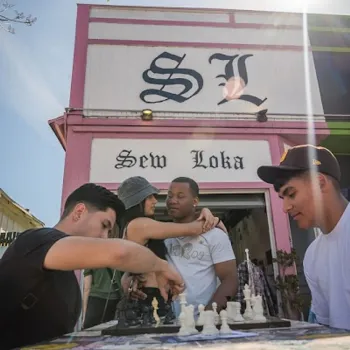 Four people interact outside a pink and white building labeled "Sew Loka"; two play chess, while two others stand close behind them. Stickers decorate the chess table.