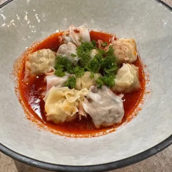 A bowl of dumplings in red chili oil sauce, topped with chopped green onions.