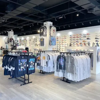 Clothing store interior with racks of graphic t-shirts in the foreground and a display wall of sneakers in the background under bright lighting.