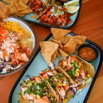 Three plates of tacos with tortilla chips, salsa, and lime wedges, served on a wooden table. The tacos feature fish, vegetables, and garnishes.
