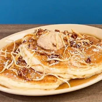 A plate of pancakes topped with a scoop of butter, powdered sugar, syrup, candied pecans, and thin strips of fried dough sits on a wooden table against a blue background.