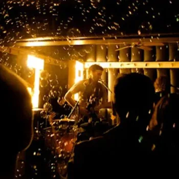 Musician performing on stage in a dimly lit venue with an audience in the foreground and bubbles floating in the air.