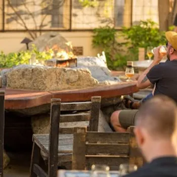 People sit around a large outdoor stone fire pit at a patio, drinking beverages and conversing on a sunny day.