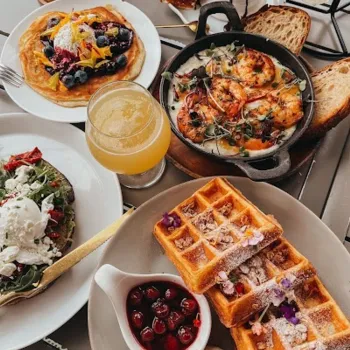 A table set with waffles, pancakes topped with berries, shrimp skillet, avocado toast, cherry sauce, whipped cream, a pastry, coffee, and orange juice.