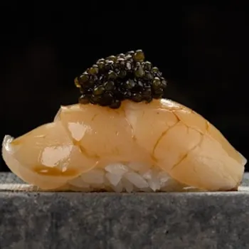 A piece of sushi with a slice of fish topped with black caviar sits on a stone surface against a dark background.